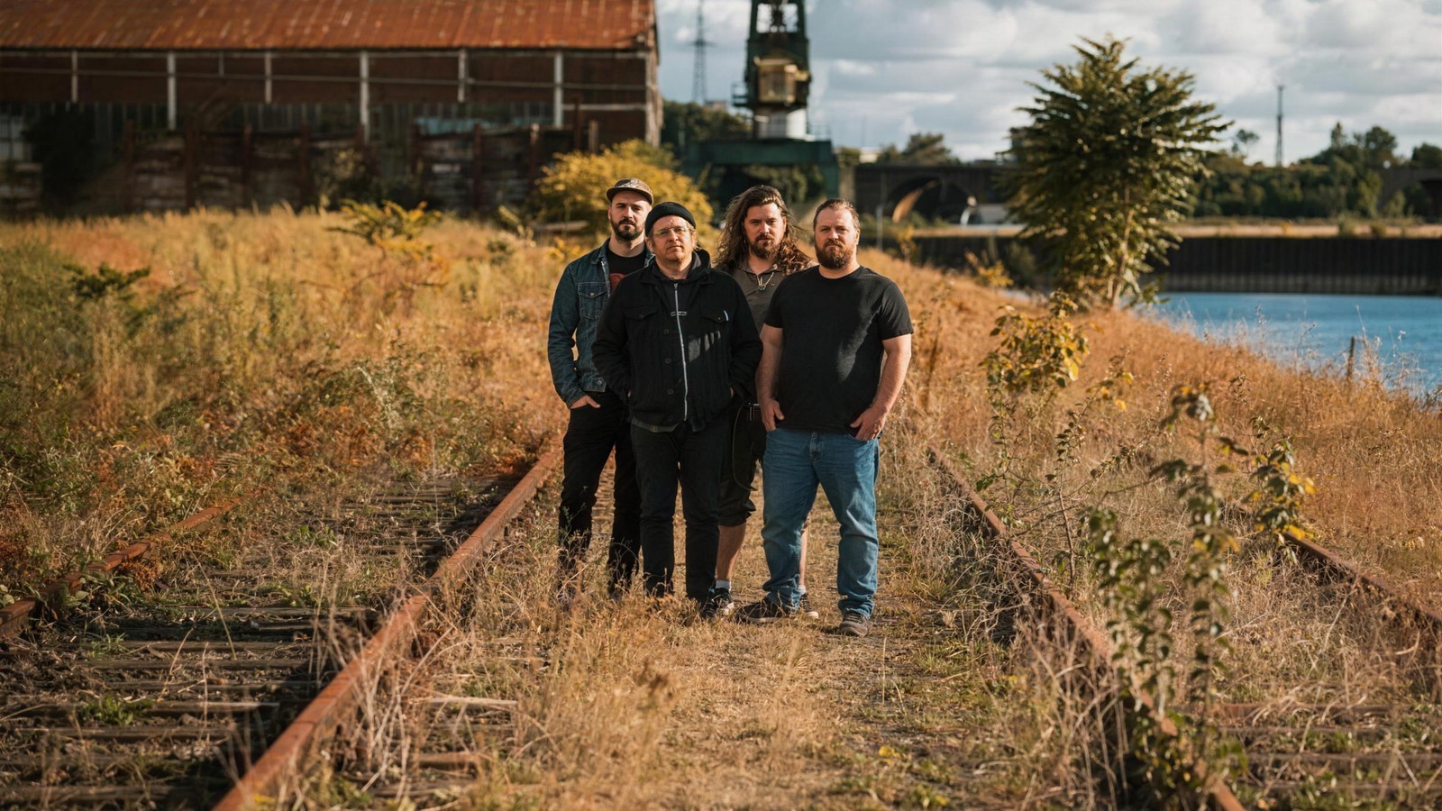 The four members of Kardashev standing on disused railway tracks overgrown with dry grass beside a river, an old dock crane and a brick warehouse behind them