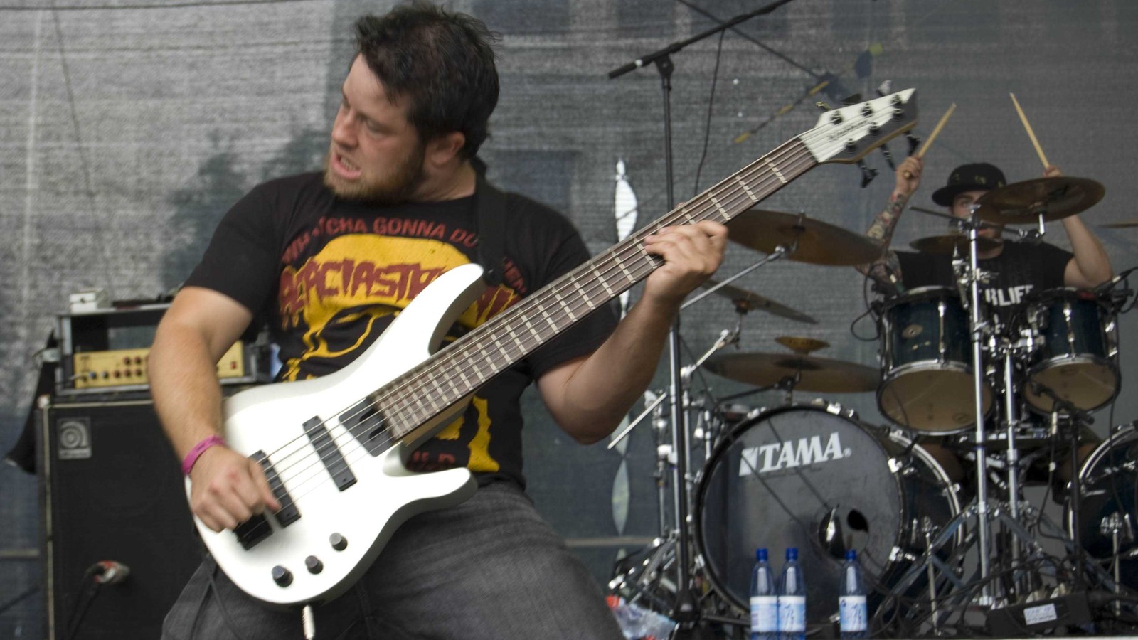 All Shall Perish bassist Mike Tiner leaning back with a white six-string bass on an open-air stage, the drummer in a cap raising his sticks behind him, 2008