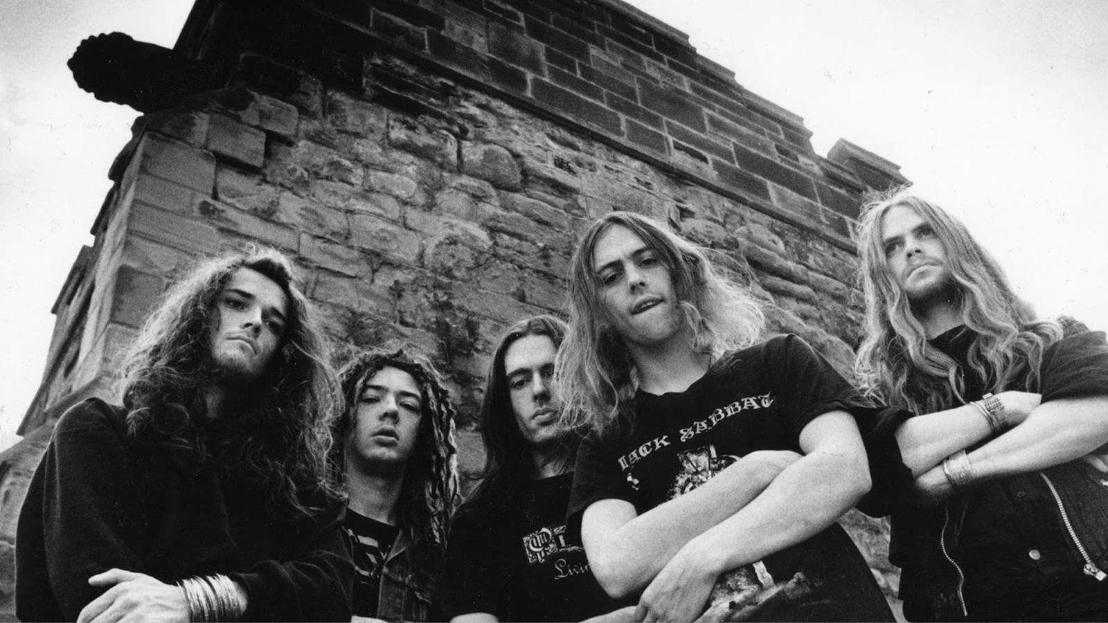 The five members of the early Cathedral line-up photographed from below against a sandstone tower, in black and white