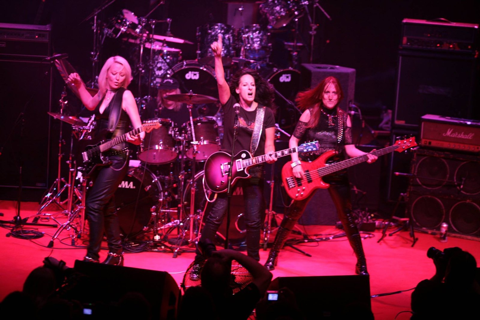 Girlschool performing live on stage, four members playing guitars and bass under red stage lighting