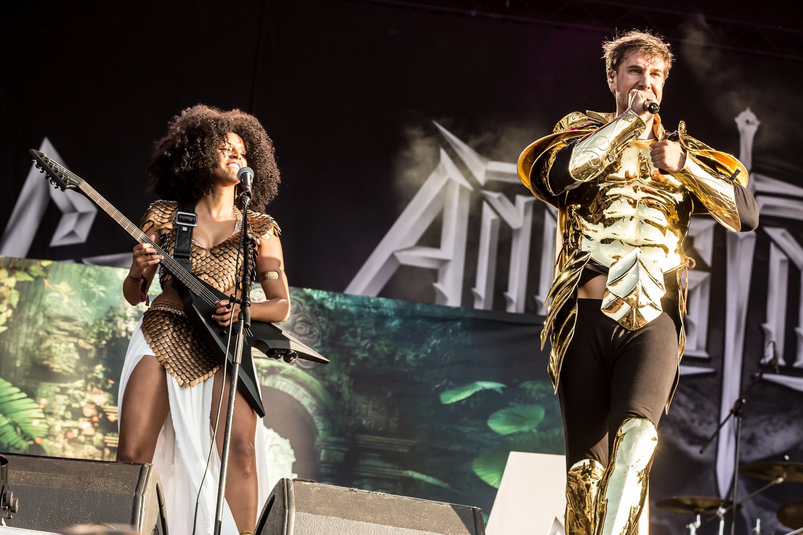 Two members of Angus McSix on stage at Rockharz Open Air 2023: a guitarist smiles beside a microphone stand while the singer in gold armour sings into a handheld microphone.