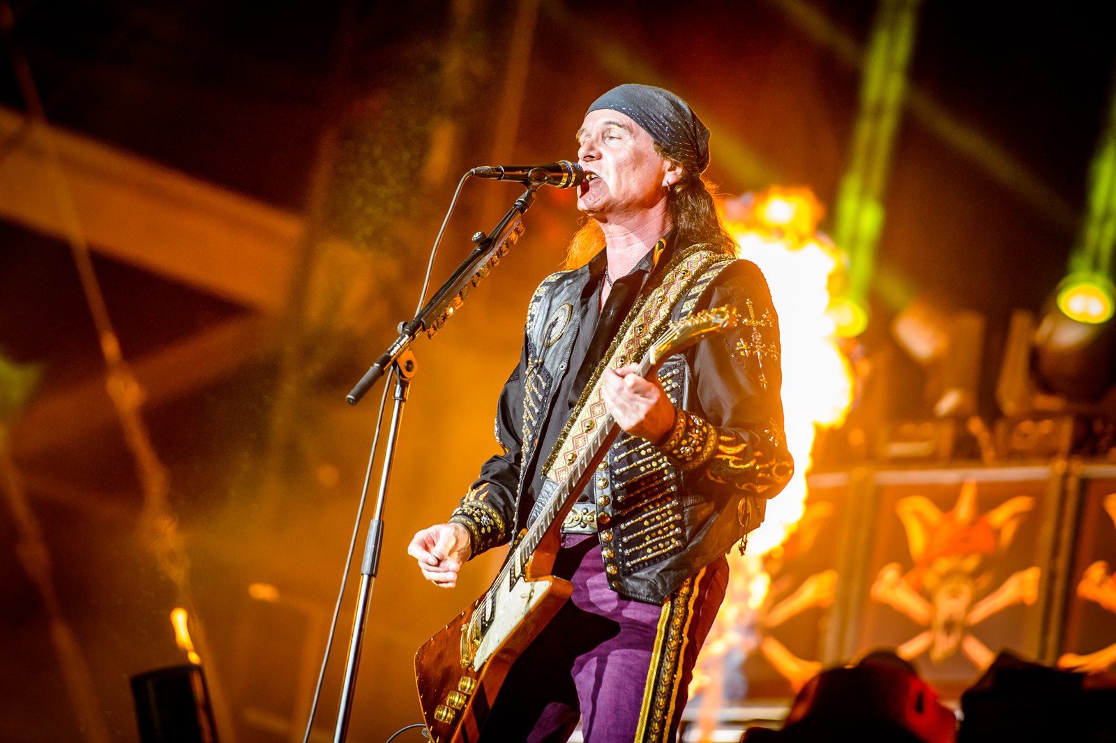 A singing guitarist of Running Wild at a microphone in a bandana and a studded black jacket with gold trim, a burst of flame behind him and the band's skull-and-crossbones backdrop at the right edge of the stage