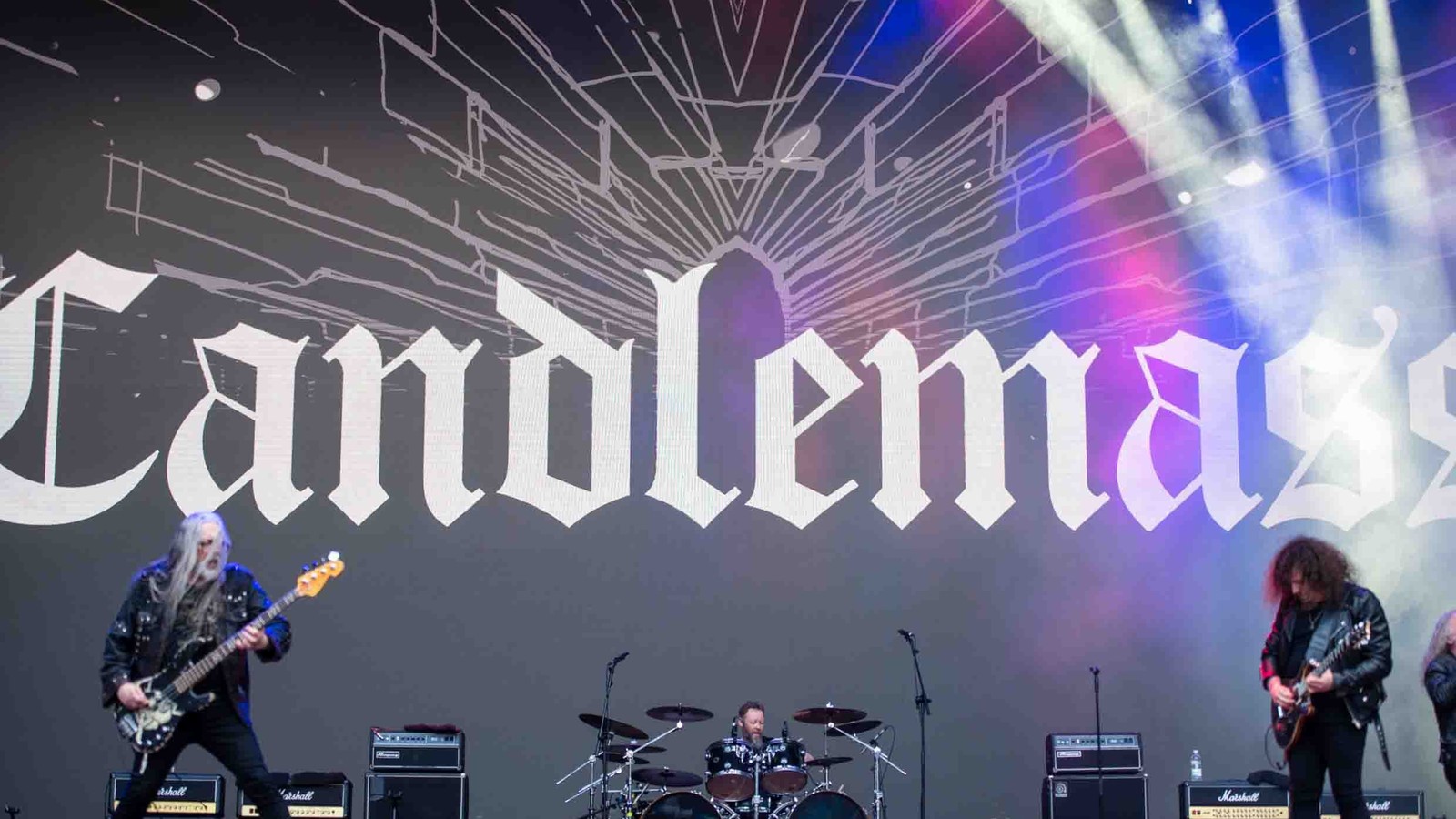 Candlemass performing on a large outdoor festival stage, the band name in white gothic letters on the backdrop