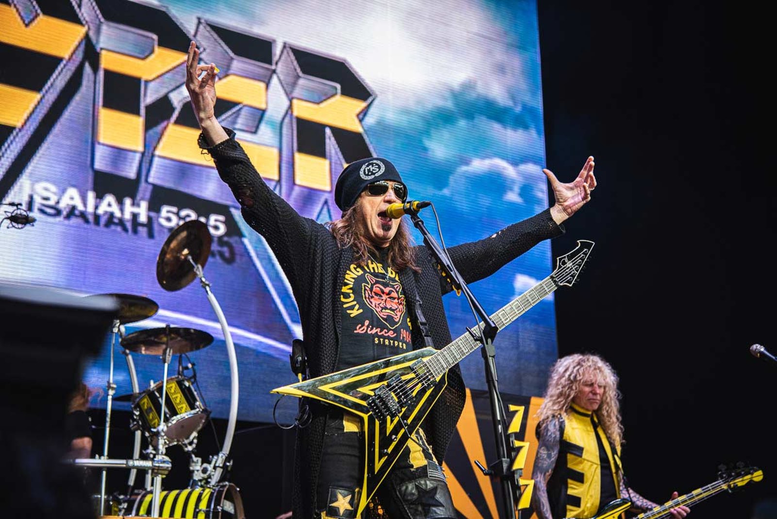 Michael Sweet of Stryper with his arms raised at the M3 festival in May 2022, the band backdrop reading Isaiah 53:5 behind him