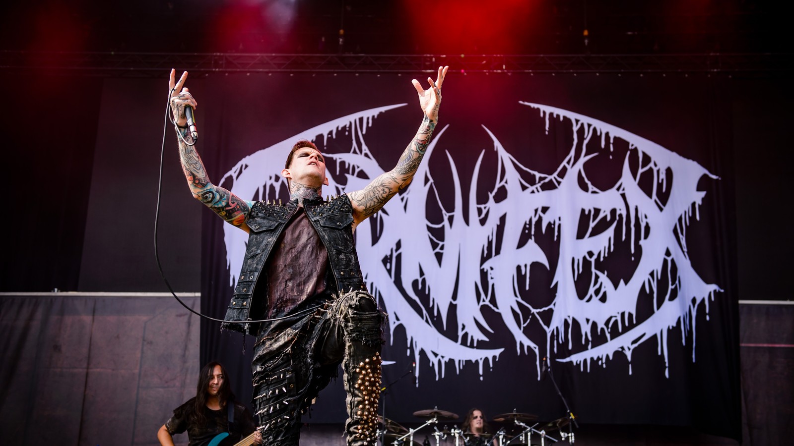 Carnifex's vocalist at Summer Breeze 2018, arms raised and one leg up, in front of the band's large white dripping logo banner, with the bassist and drummer behind.