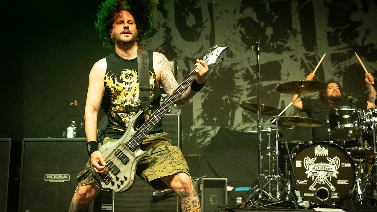 Suicide Silence's bassist playing, hair flying, in front of the band's ink-spattered logo backdrop at Milwaukee Metal Fest