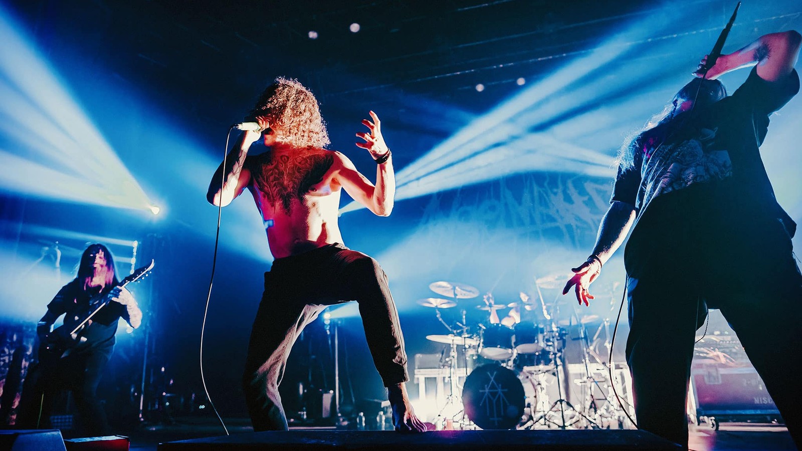AngelMaker on stage at L'Olympia in Montreal, the shirtless vocalist crouched on a riser mid-scream, a second vocalist at right and a guitarist at left under blue light