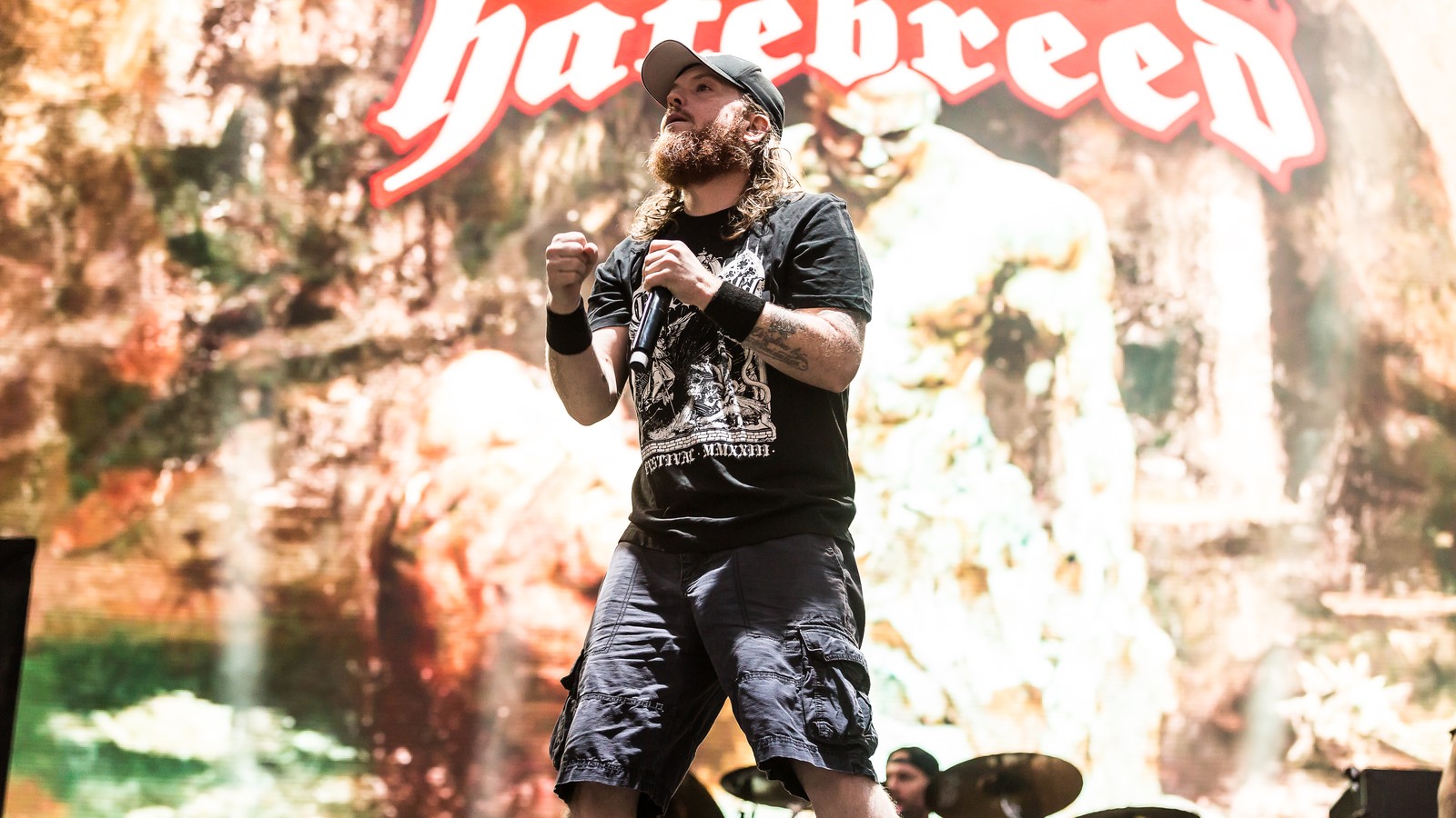 Hatebreed singer Jamey Jasta mid-song on the With Full Force 2023 stage, fist clenched and microphone in hand, the band's flaming logo on the video wall behind him.