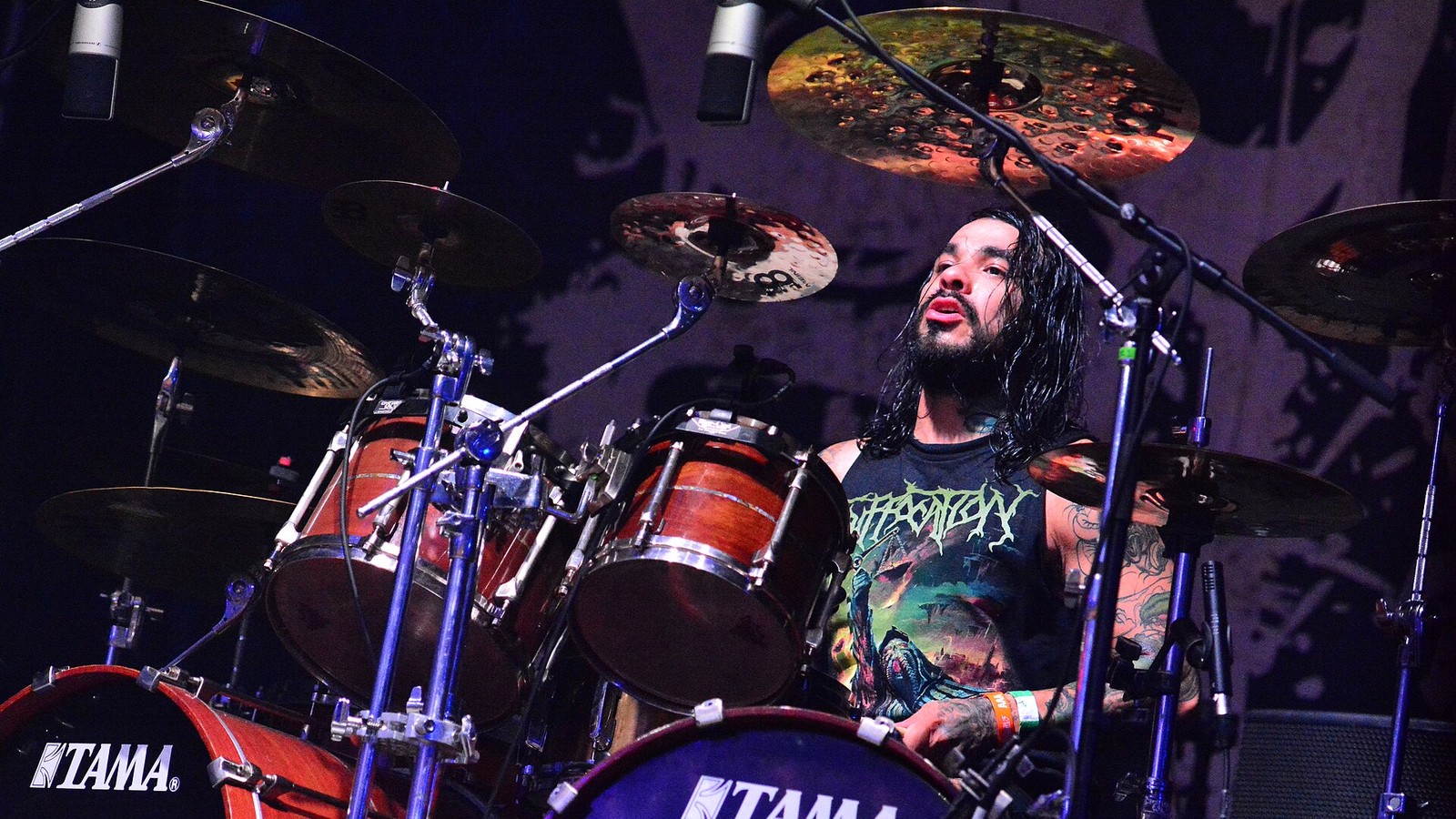Suicide Silence drummer Alex Lopez behind his kit at Wacken Open Air, hair soaked in sweat, cymbals and microphones around him and the band backdrop behind.