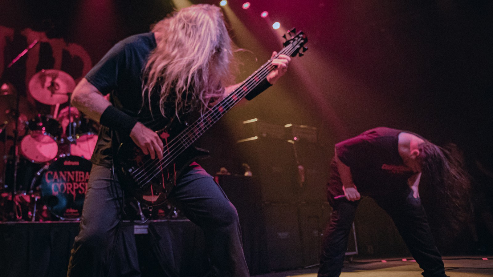 Cannibal Corpse on a red-lit stage, the bassist bent over a five-string bass in front while the vocalist headbangs bent double at right, the band's logo on the kick drum behind.
