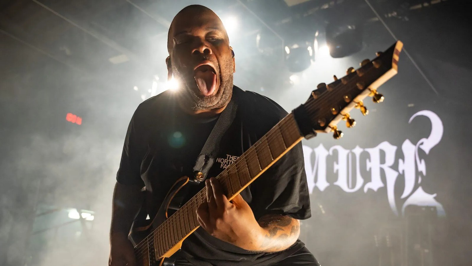 Emmure guitarist leaning toward the camera with an extended-range guitar angled across the frame, headstock and fretboard in focus against stage smoke and the band's logo, Max Watts, Melbourne, 2024