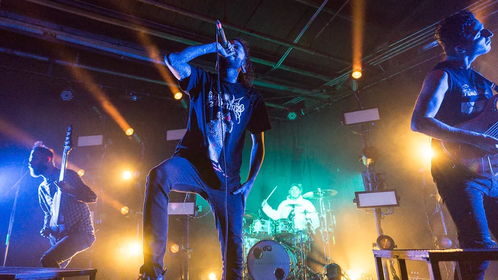 Chelsea Grin vocalist Tom Barber with the microphone at his mouth under orange and teal stage light, guitarist, bassist and drummer behind him.
