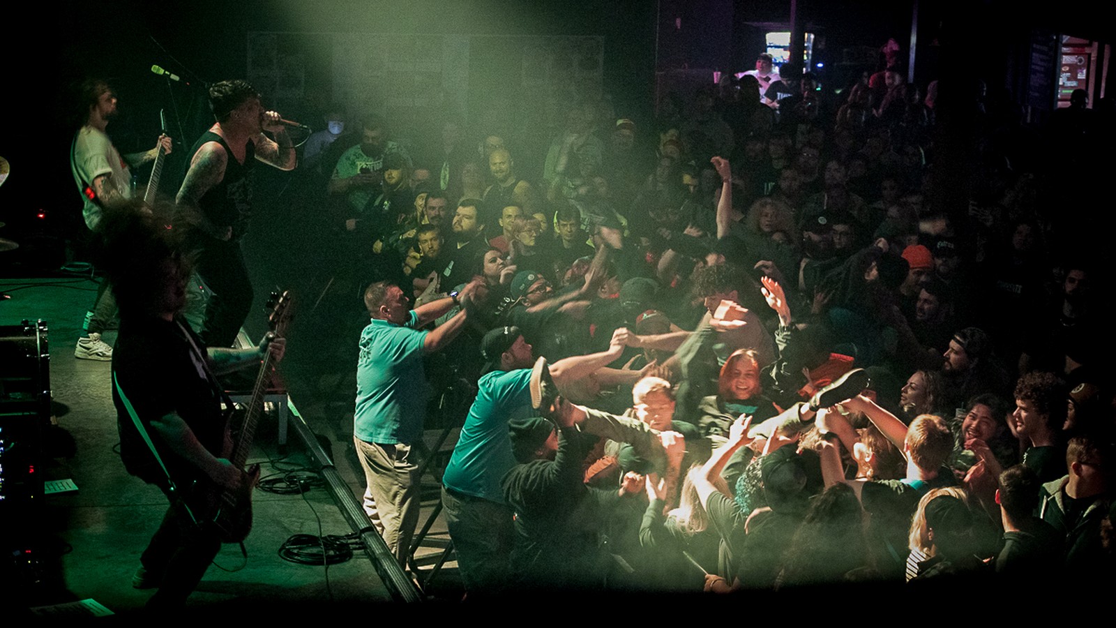 Bodysnatcher on stage at Red Flag in St. Louis in November 2023, vocalist and guitarists at the left while security staff in teal shirts pull crowd surfers over the barrier from a packed, green-lit pit.