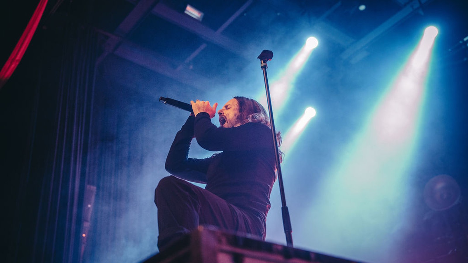 Fit for a King vocalist Ryan Kirby kneeling on a stage monitor and singing into the microphone under blue and teal beams at The Pageant, St. Louis, 2025.