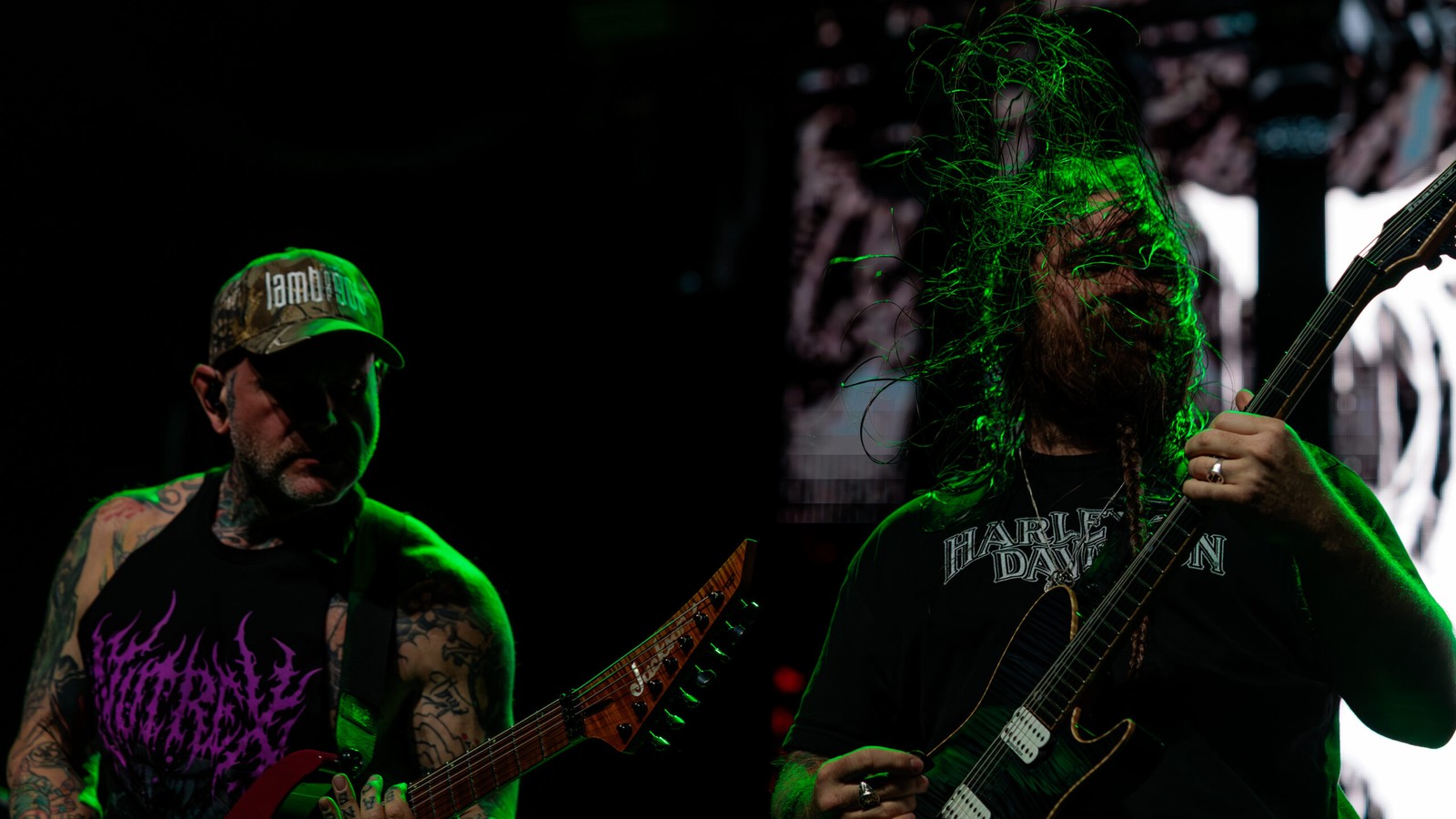 Two Fit for an Autopsy guitarists playing side by side on stage at the WAMU Theater in Seattle, one in a cap and one with long hair thrown forward, under green light