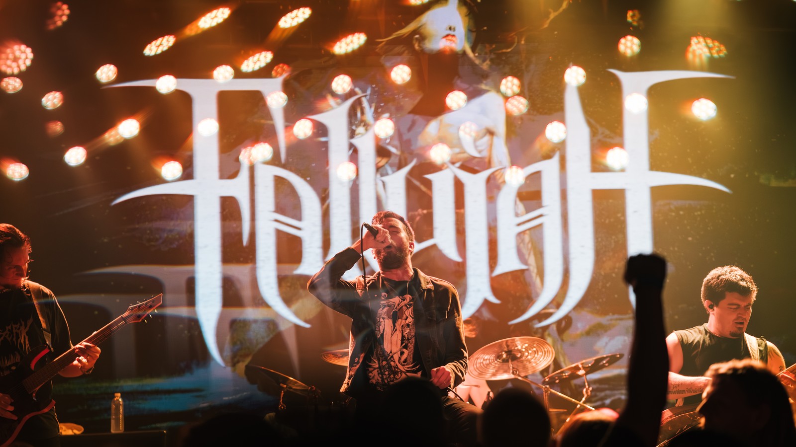Fallujah vocalist at the microphone in front of the band's logo on the stage backdrop, a guitarist at left and the drummer at right, a fist raised in the crowd, Goldfield, Roseville, July 2023