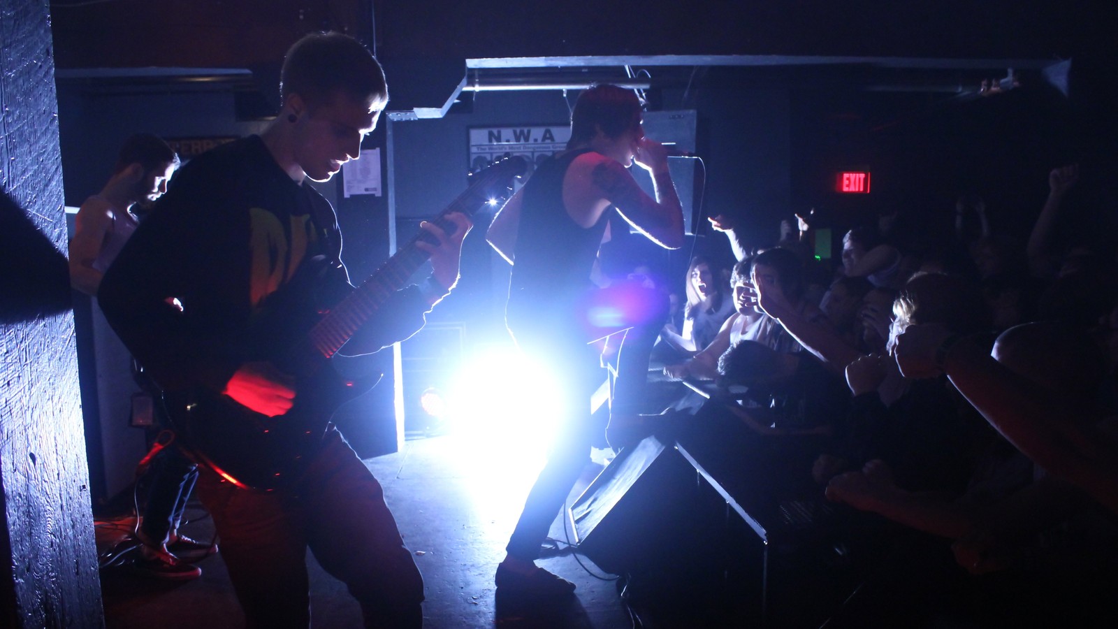 Chelsea Grin's vocalist at the microphone in a backlit club, one hand raised, a guitarist in the foreground and the front row reaching toward the stage.