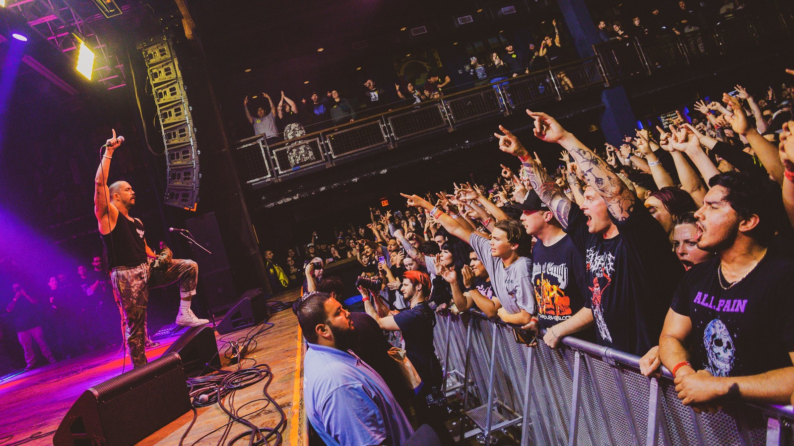 Kublai Khan TX vocalist on stage with one arm raised, facing a crowd pressed against the barricade with hands and fingers pointing back at him, a balcony full of fans above.