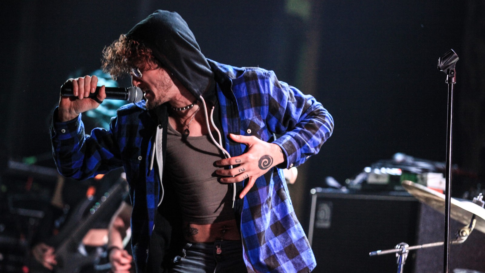 Tallah's vocalist in a hood and a blue flannel shirt screaming into the microphone, leaning forward on a club stage with the bassist behind him