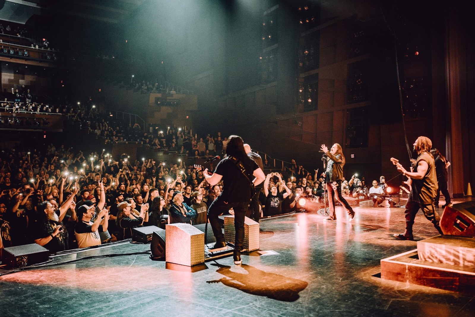 Dream Theater performing live on stage, the singer reaching toward the crowd as the guitarist and bassist play behind him under red and white stage lights