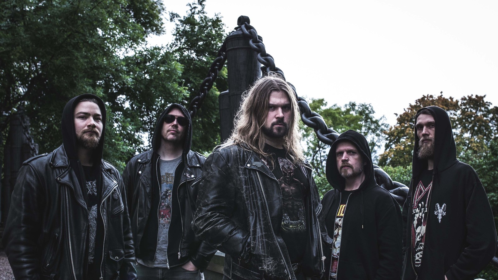The five members of Hooded Menace in leather jackets and hoods standing in front of a monument hung with giant iron chains