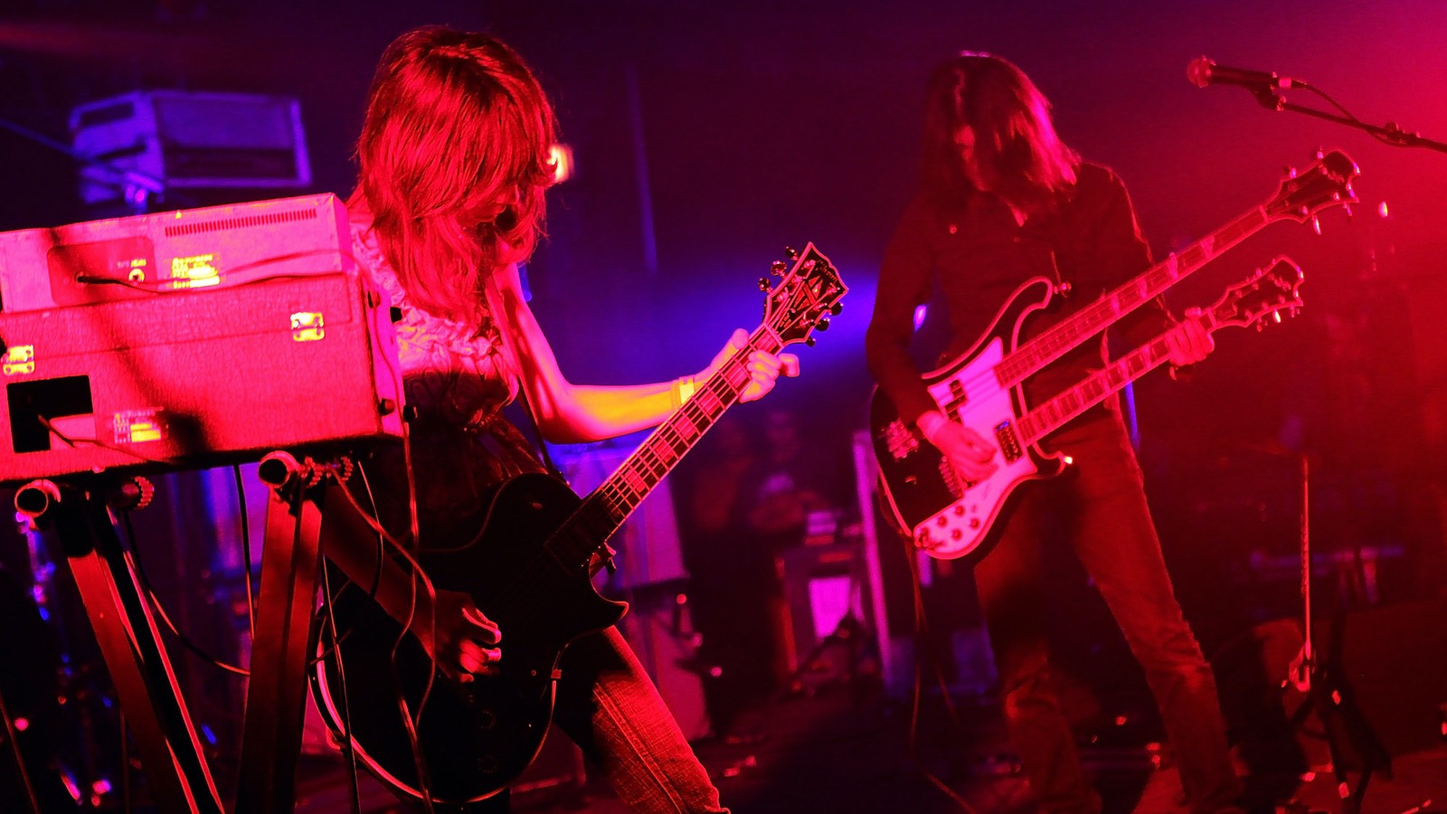 Boris guitarist Wata playing beside an amplifier while Takeshi plays a double-neck guitar and bass, both lit pink and purple
