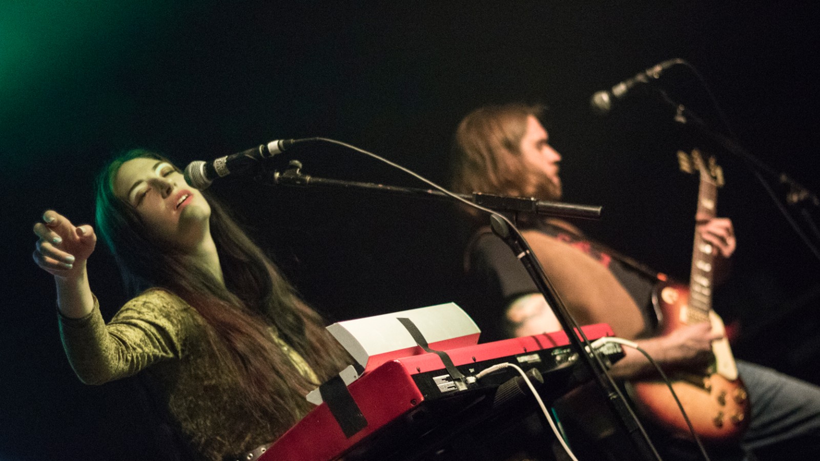 Blood Ceremony singer Alia O'Brien in a crushed velvet dress singing behind a red organ, the guitarist playing behind her