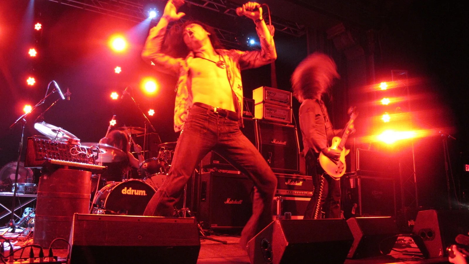 Church of Misery's singer leaping with both arms raised under red stage light, the guitarist headbanging beside a Marshall stack