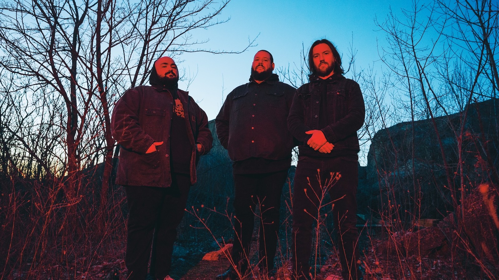 The three members of Primitive Man standing among bare trees and dry grass at dusk, lit red against a blue sky