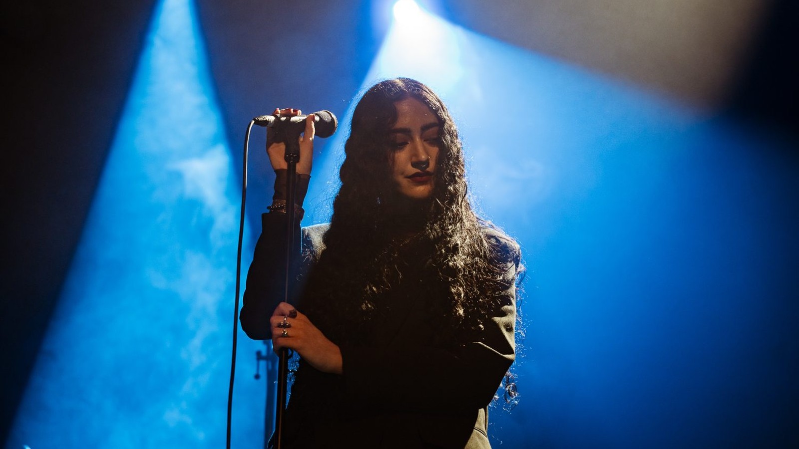 The singer of Messa with long dark hair at the microphone, lit from behind by blue spotlights