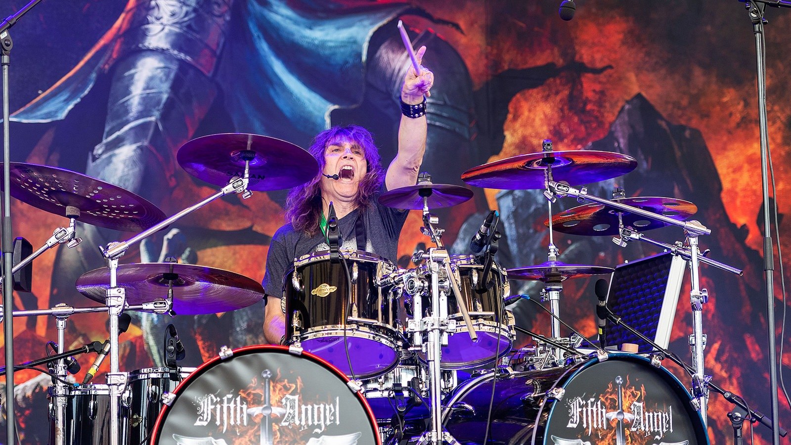 The drummer of Fifth Angel with a stick raised behind a kit whose two bass drums carry the band name, a painted knight and fire on the backdrop