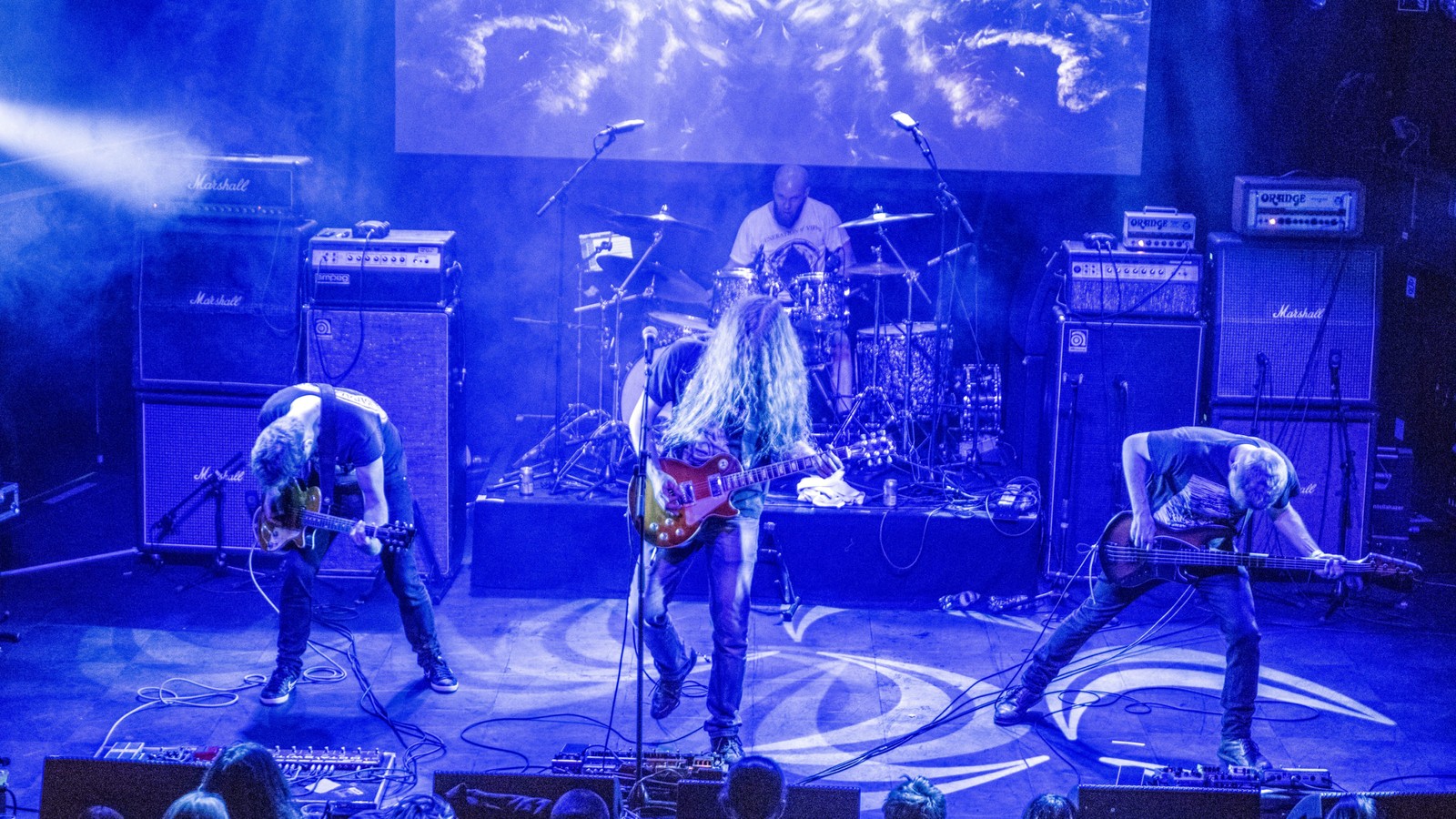 Ahab performing on a concert stage in deep blue light, guitarists and bassist lined up in front of their backdrop