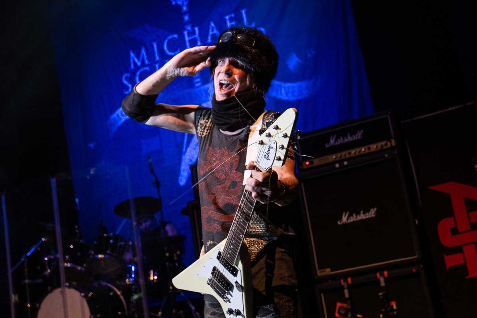 Michael Schenker on stage with his black and white Flying V, one hand raised to the crowd, a wall of amplifiers behind him