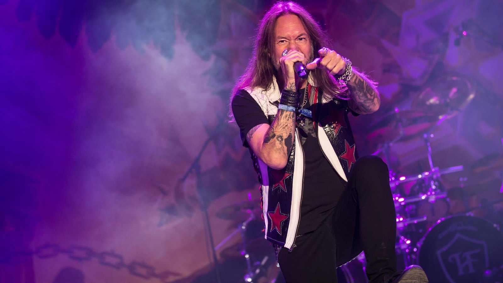 HammerFall singer Joacim Cans points into the crowd from the front of the stage at Summer Breeze in 2019, the band's shield emblem on the drum kit behind him