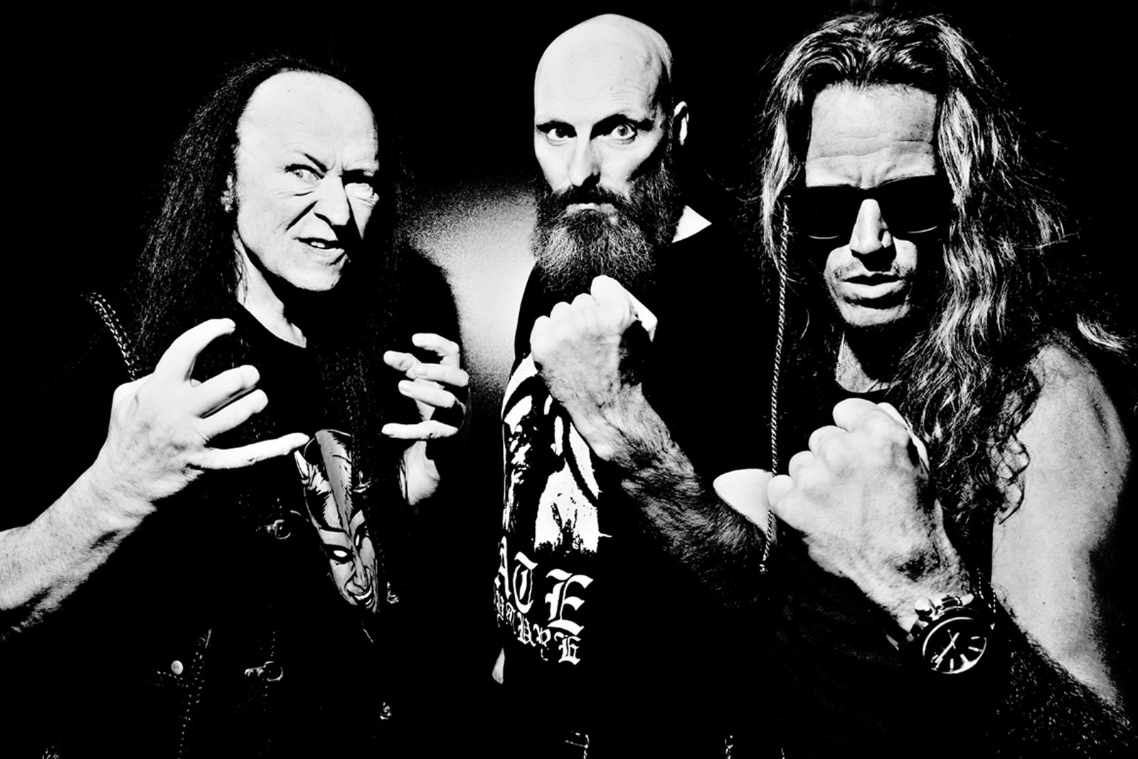 Venom band portrait in black and white, three members in black band t-shirts and leather making devil-horn and fist gestures