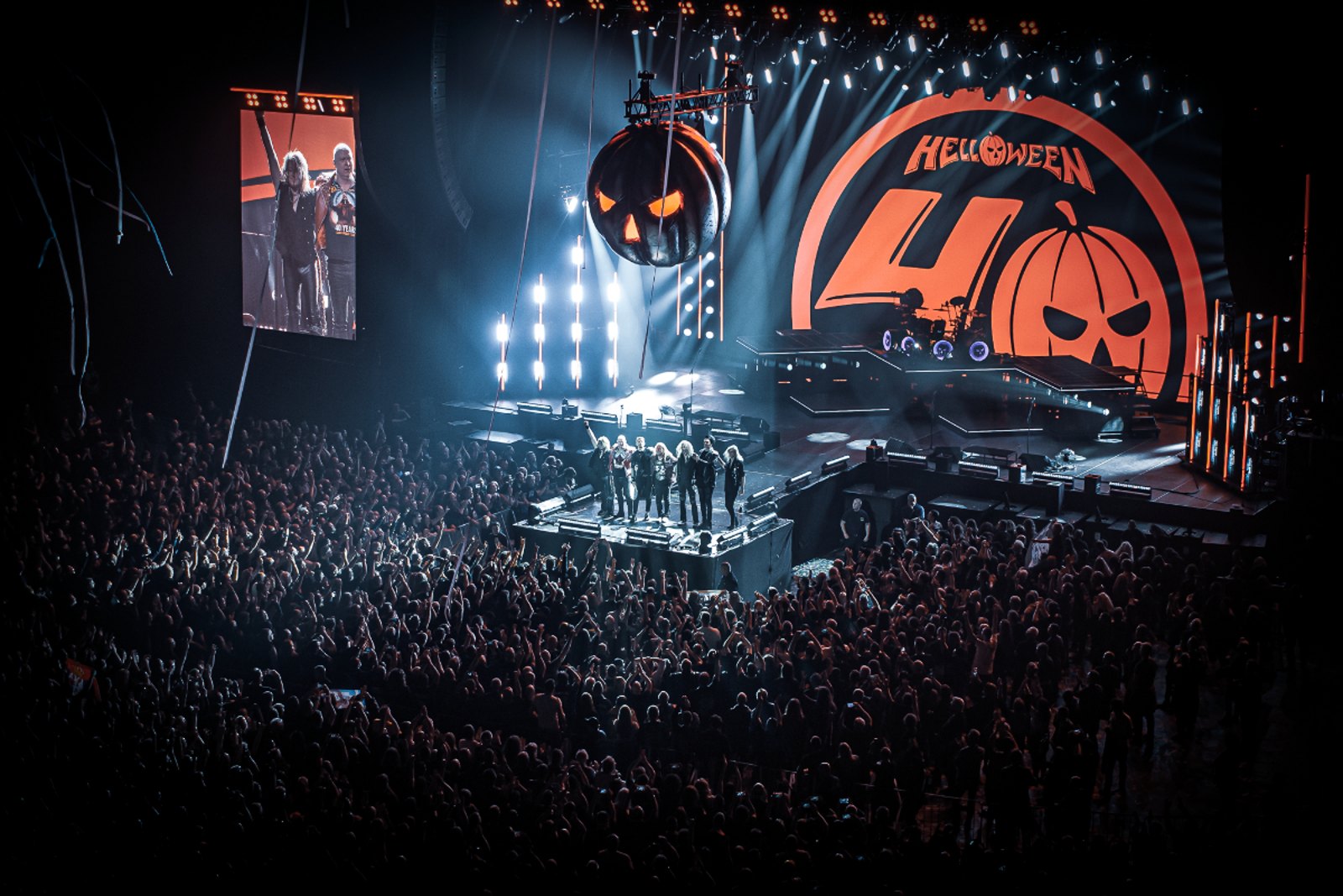 Helloween on stage in Budapest beneath a hanging carved pumpkin and a backdrop whose forty is closed by a jack o lantern face