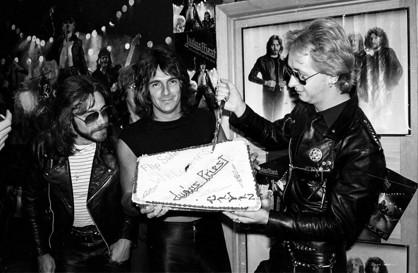 Judas Priest members in leather cutting a cake at a 1979 record store event welcoming the band, tour posters on the wall behind them