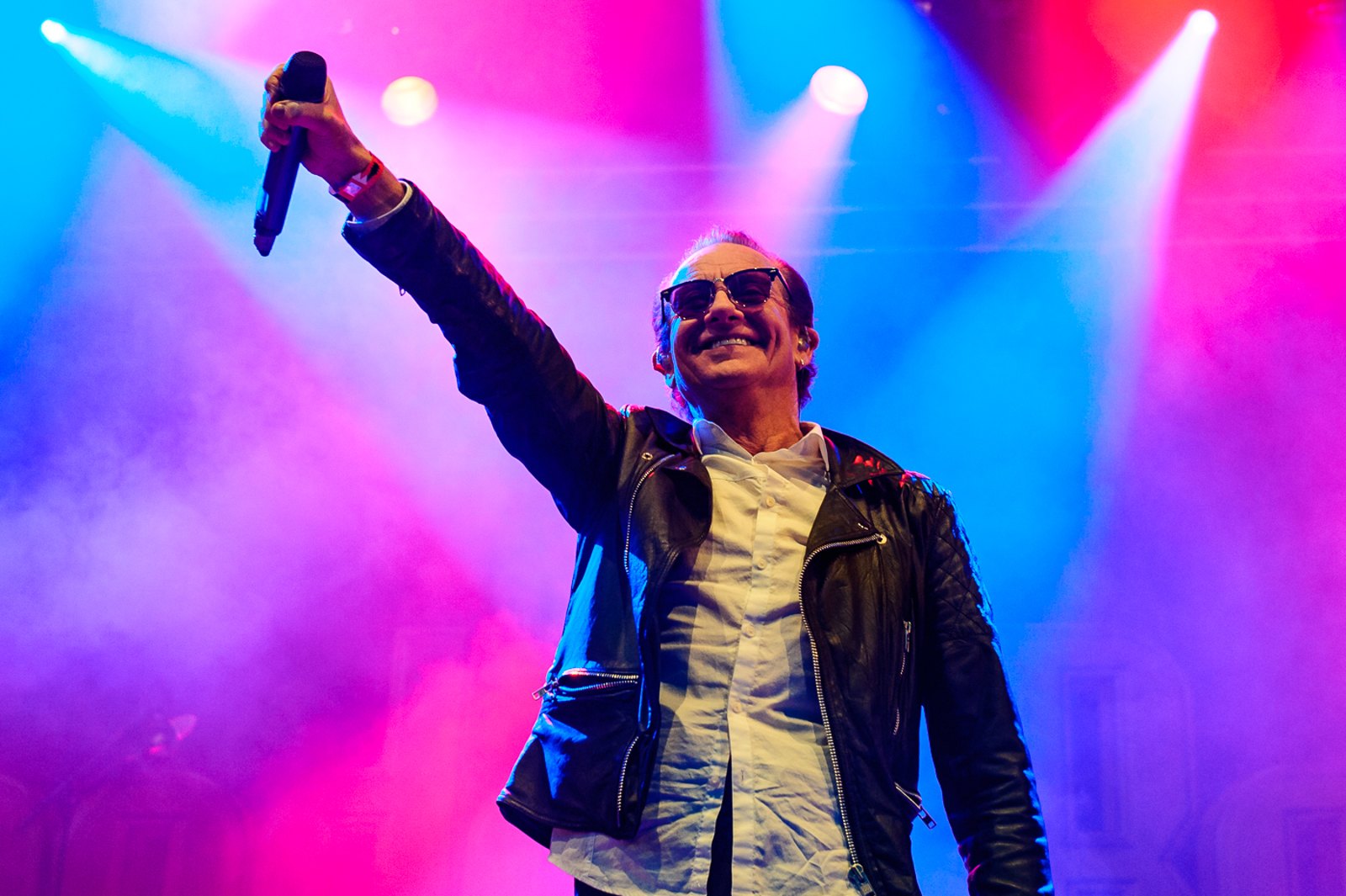 Graham Bonnet performing live at Sweden Rock Festival, 2016, raising the microphone with one arm as stage lights sweep blue and pink behind him