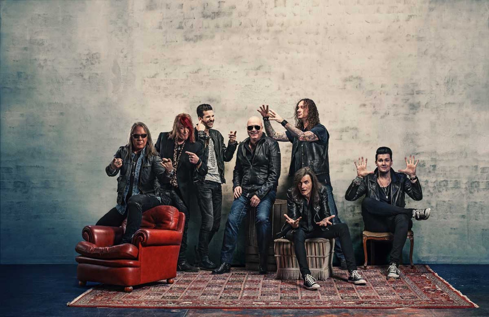 The seven members of the reunited Helloween line-up posed together in a studio, some seated on a red armchair and a crate, others standing behind them