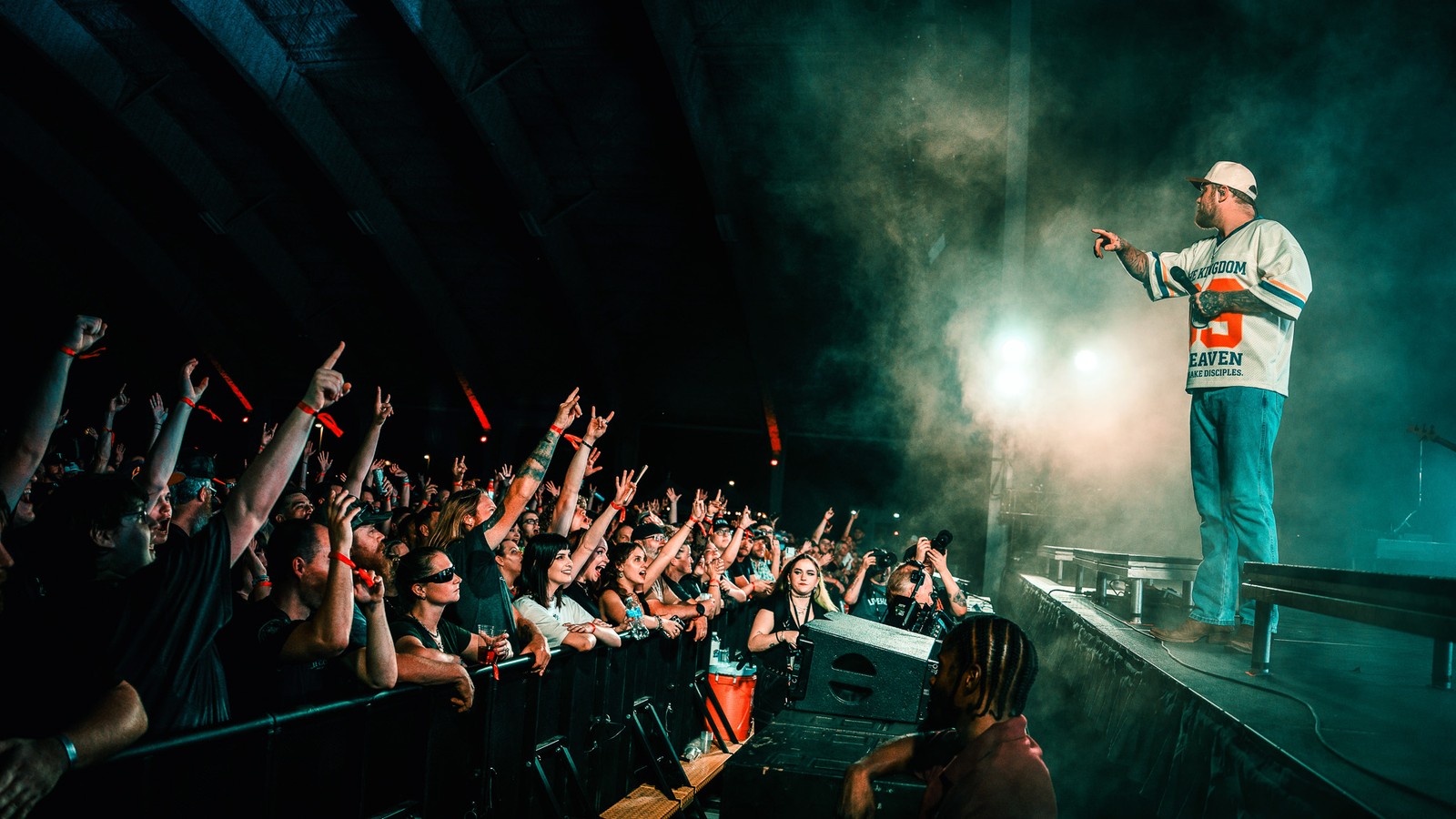 Asking Alexandria singer Danny Worsnop in a white jersey and cap on a raised stage ramp, pointing toward a crowd of raised fists in stage haze