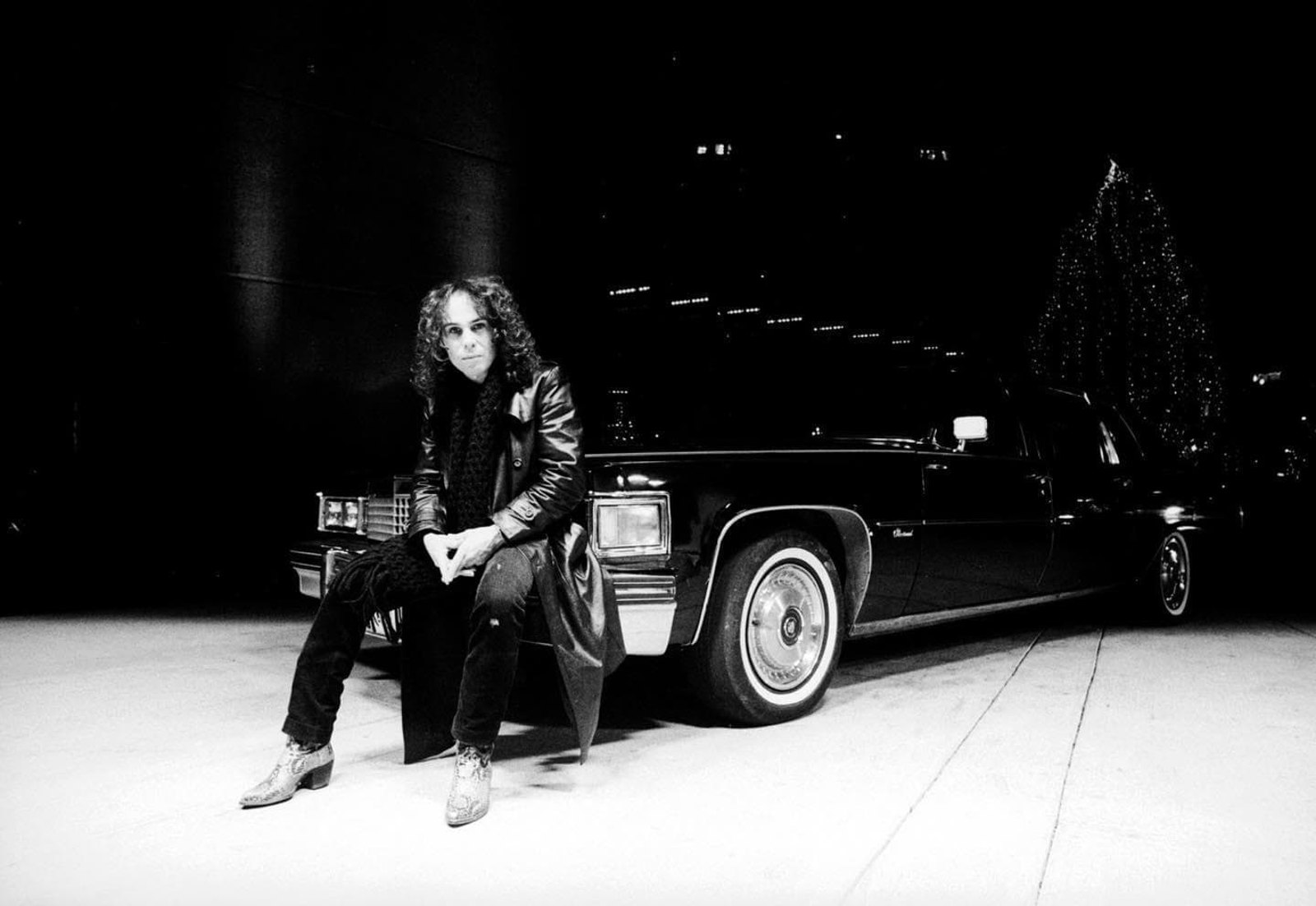Ronnie James Dio in a black and white photograph, sitting on the front wing of a limousine at night with a lit tree behind him
