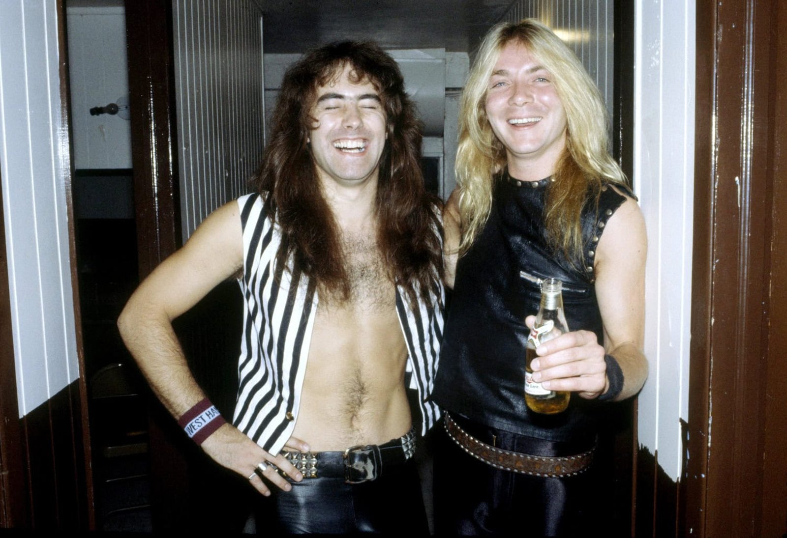 Two members of Iron Maiden laughing backstage in 1982, one wearing a black and white striped waistcoat and holding a bottle