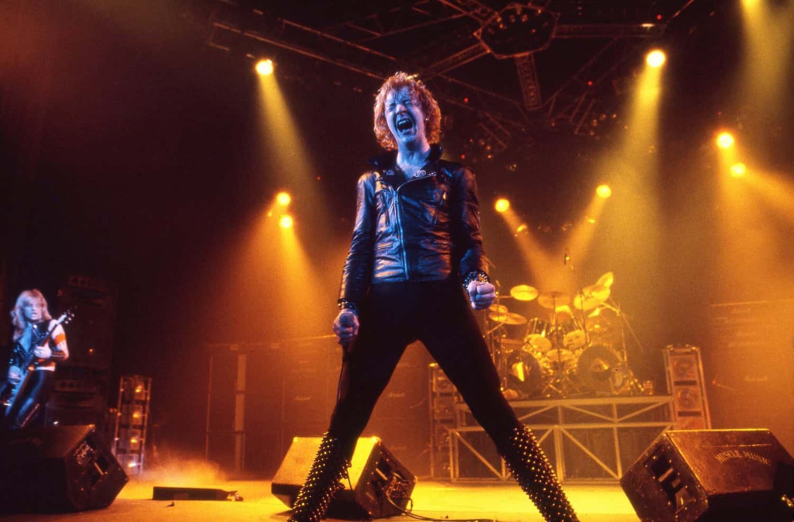 Rob Halford of Judas Priest singing into a hand-held microphone on stage, studded leather jacket and raised gauntlet, guitarist beside him