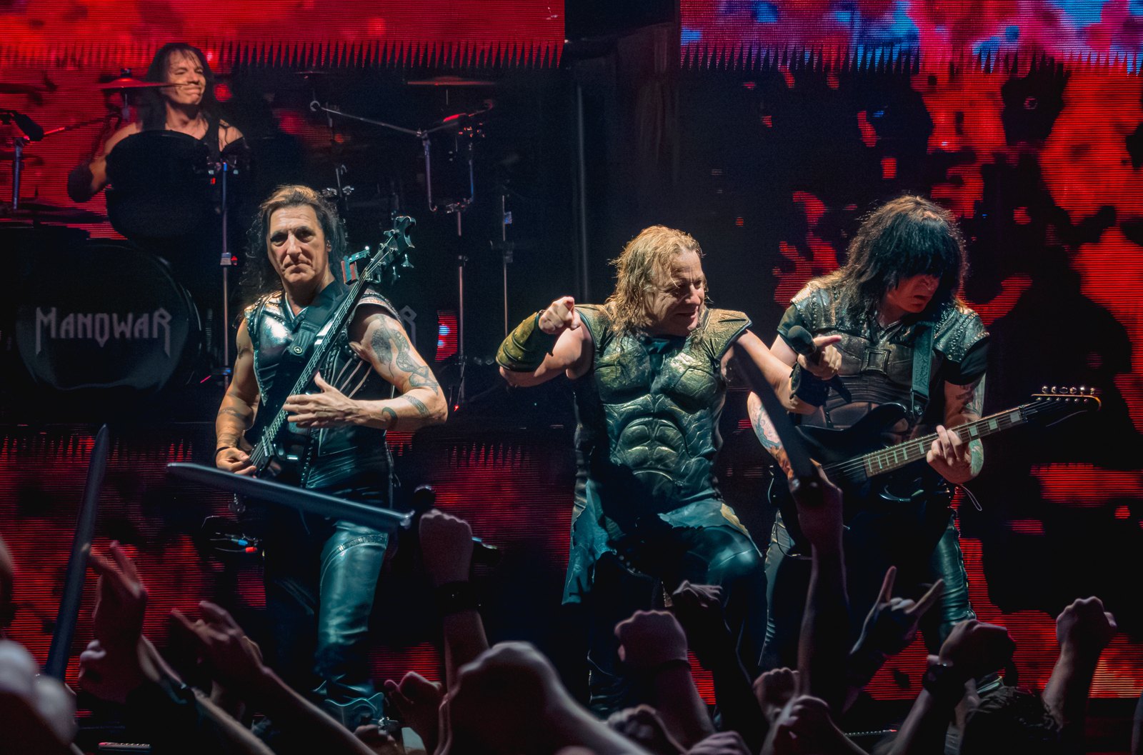 Manowar on stage at the O2 Academy Brixton, London: bassist Joey DeMaio, singer Eric Adams pointing at the crowd and a guitarist, with raised fists below and the drummer behind.