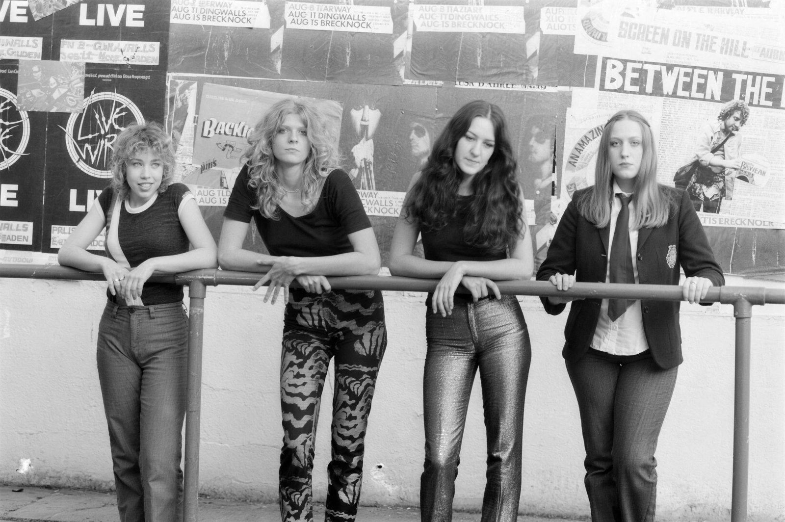 Girlschool's four original members leaning on a railing in front of gig posters for Dingwalls and Brecknock, London, around 1980