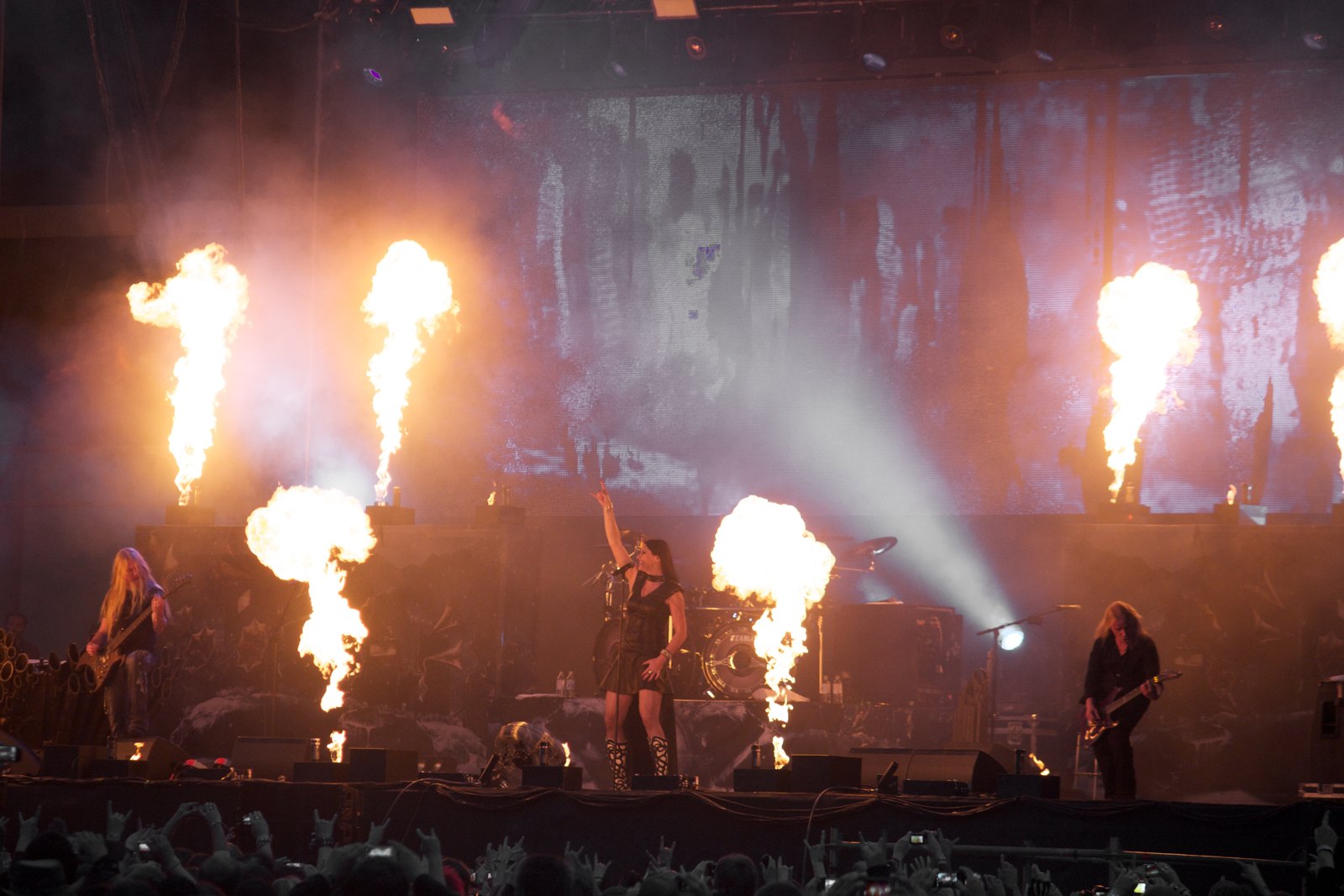 Nightwish on the M'era Luna Festival stage in 2013: Floor Jansen with an arm raised at centre, bassist Marco Hietala and guitarist Emppu Vuorinen at the sides, flame jets going off across the stage front.