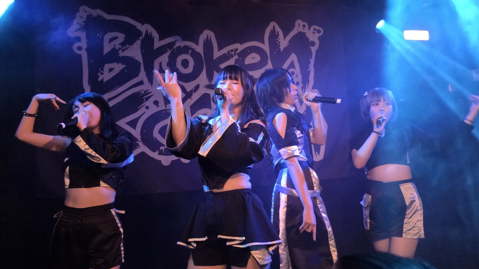 Broken By The Scream performing at Bitterzoet in Amsterdam, four vocalists in black and white stage outfits at their microphones under blue stage light, with the band's logo banner behind them.