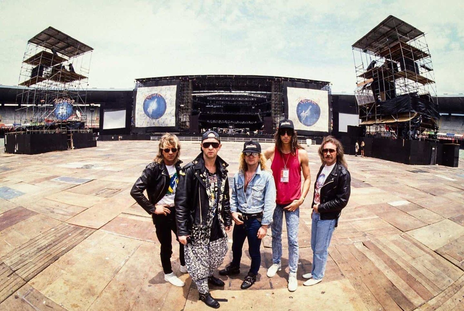 The five members of Judas Priest standing on the empty floor of a stadium in front of the stage, tour banners carrying the Painkiller sleeve art behind them