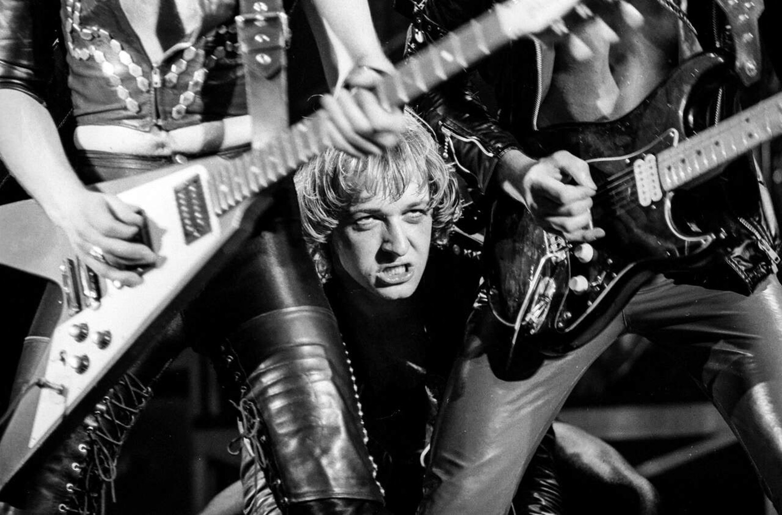 Rob Halford of Judas Priest singing live on stage in studded leather, arms outstretched, bassist and drummer behind him under yellow stage lights