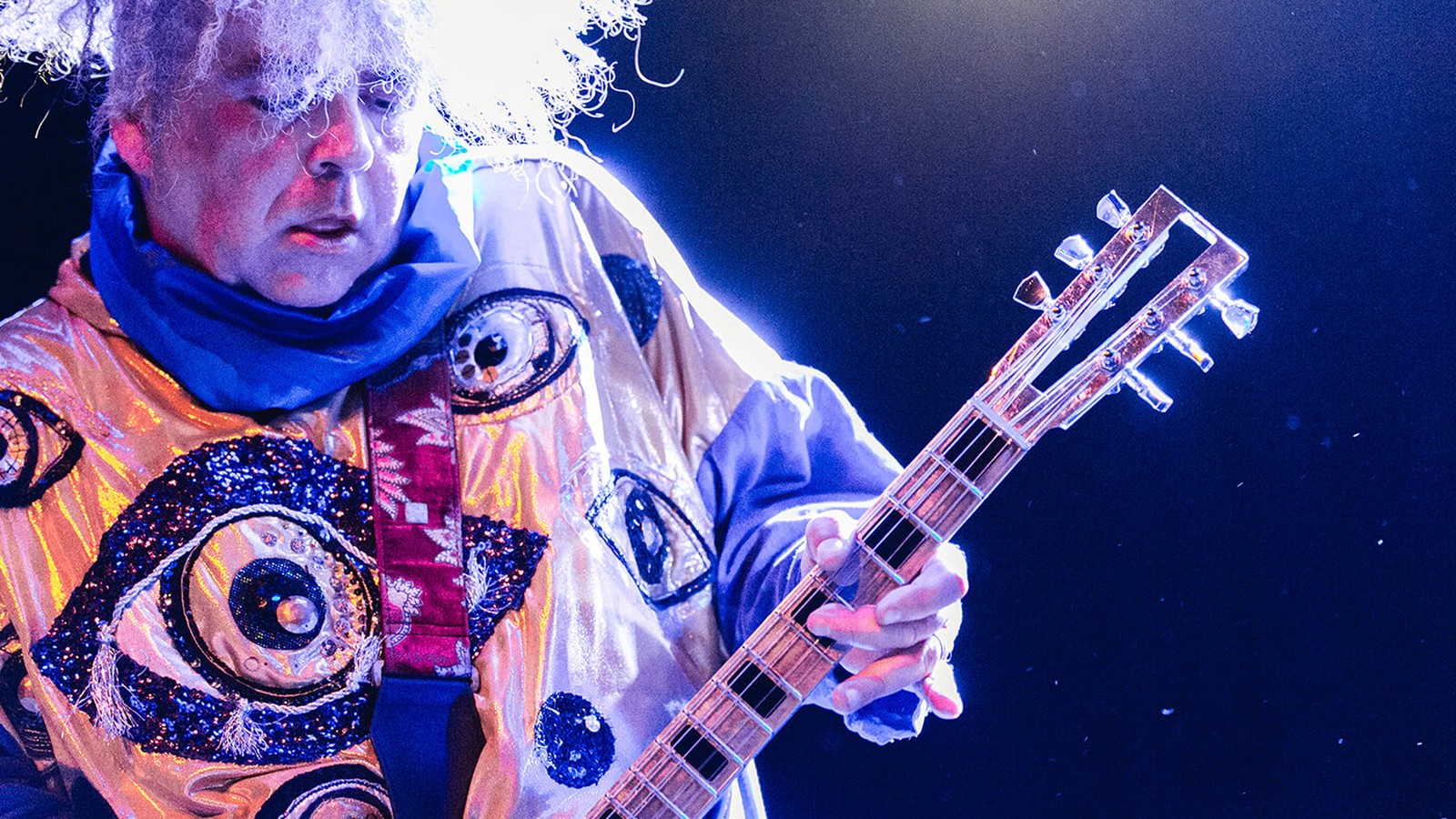 Buzz Osborne of the Melvins with his white shock of hair, playing guitar under blue light in a robe covered in eyes